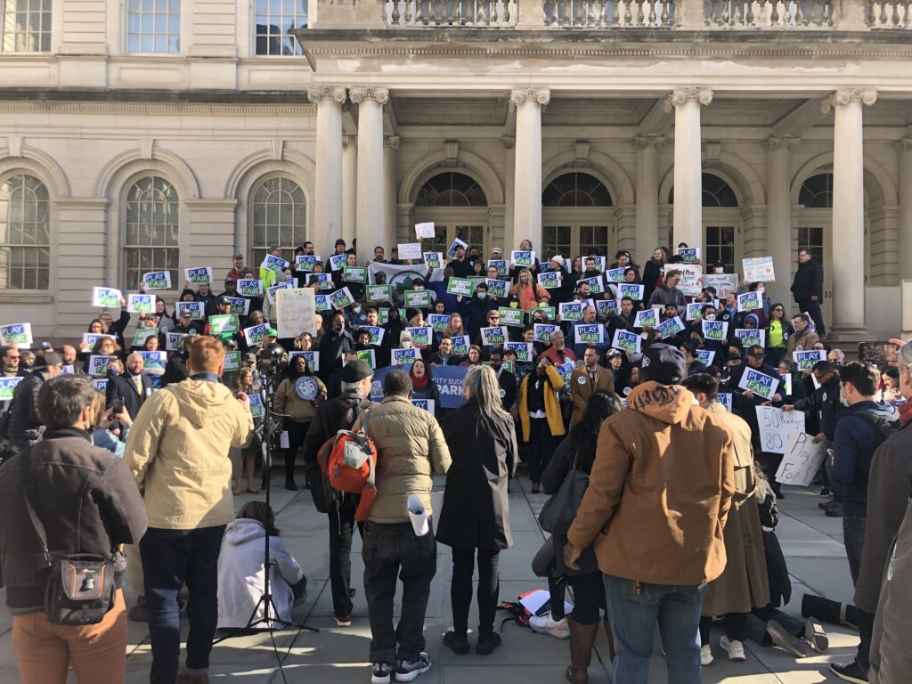 Large group of protestors holding Play Fair signs stand on steps of NYC city hall