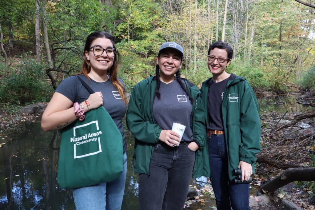 Three staff members wearing NAC attire stand in front of a pond in Central Park