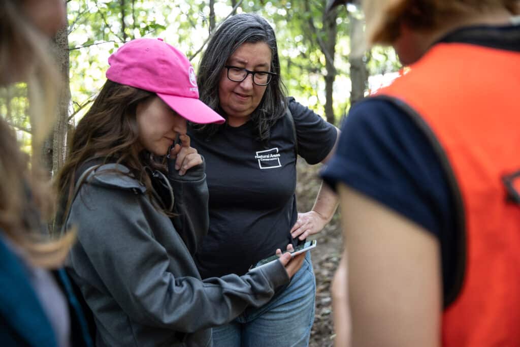 A NAC staff member and NYC Parks employee huddle over a cellphone in a forest