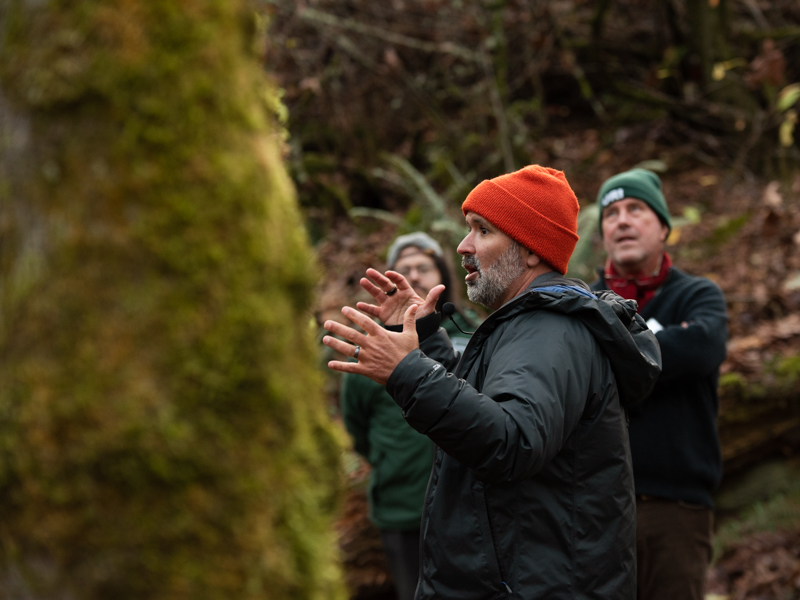 A man in an orange hat speaks to a crowd of Fic members in a forest