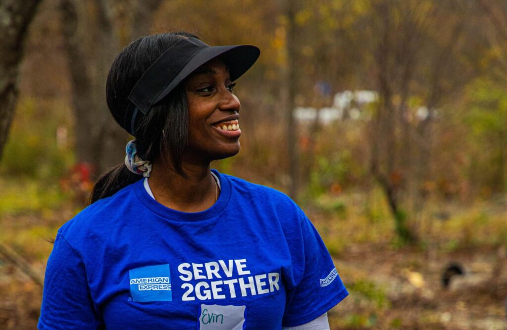 A woman holds empty tree containers wearing a shirt that reads 'AMEX SERVE 2GETHER'