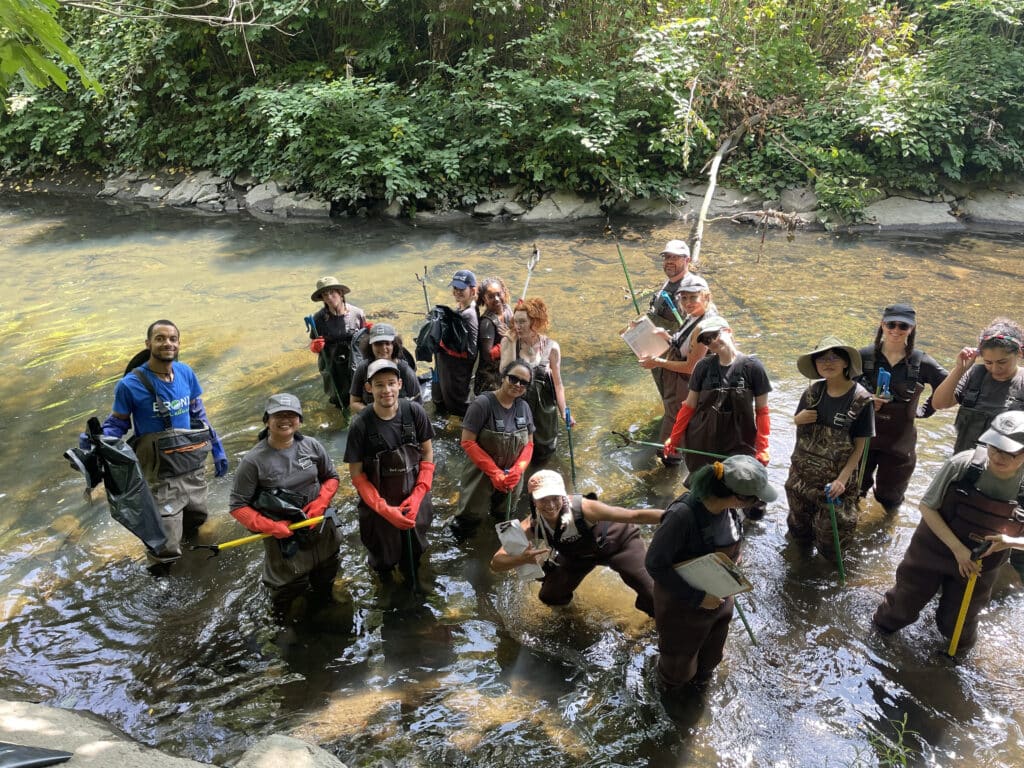 A group of interns in waders smile while holding trash bags and wearing gloves in the Bronx River