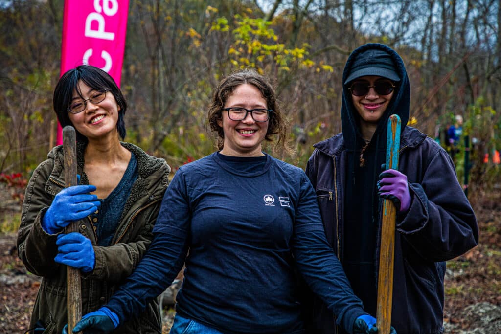 Three interns smile in a row holding shovels in a natural area