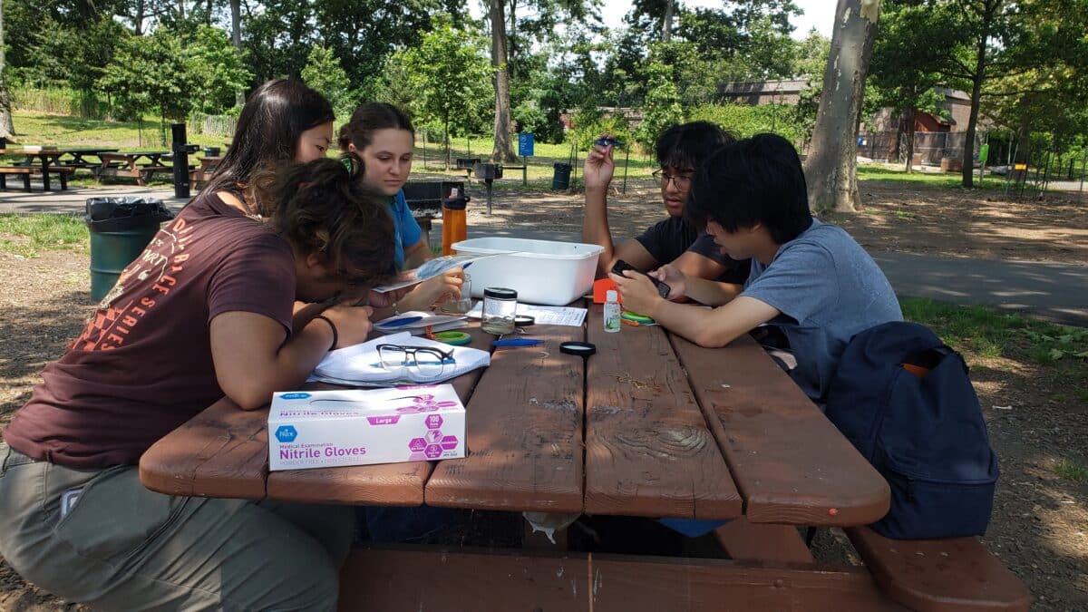 A group of five high school interns sit around a picnic table with a water sample kit